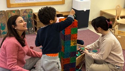 Boy building a tower with blocks while two young women sit on the floor watching.
