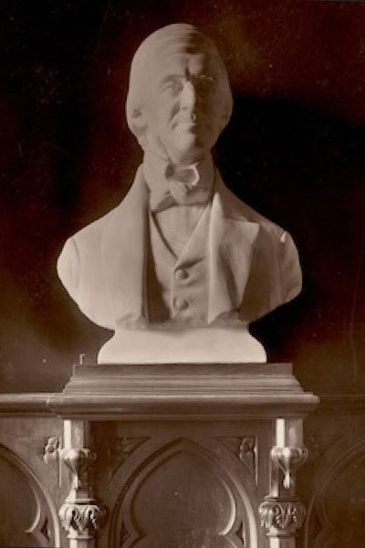An off-white bust of Emerson wearing a bow tie and suit with vest, set in a dark brown architectural setting.