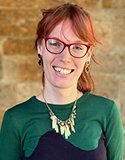 Head shot of woman with red ling hair, bands and glasses smiling.