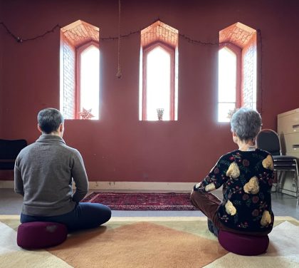 Two people sitting cross-legged, viewed from their back in a room with three narrow woindows