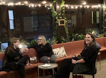 A conversation of one man and two women (one smiling) siting with glasses of wine with white twinkle lights overhead.