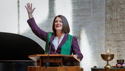 Woman with brown hair speaking from a pulpit with her hand raised, and lit copper chalice by her side