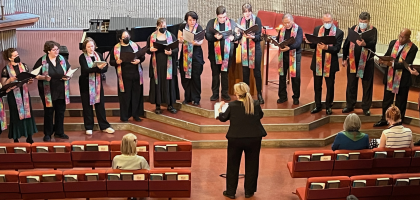 Choir with black clothes and multicolored stoles in a row in front of the church sanctuary being directed.