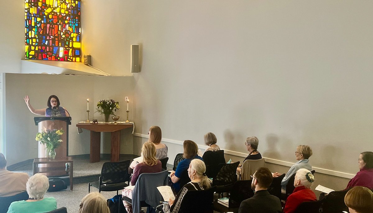 Woman at a lectern with a colorful stained glass window behind her in light-filled room and people seated listening to her talk.