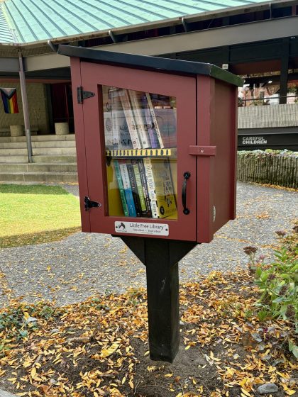 A Little Free Library (brownish box with slanted black roof) mounted on a vertical black post and contains books set in a garden.