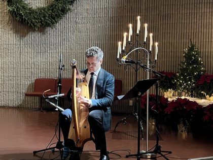 Man with grey hair and wearing a suit plays a harp with a candelabra with a wreath in the background.