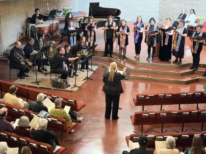 A semi circle of singers in standing on steps with musicans with instruments, being directed by a woman with blonde hair in a church.